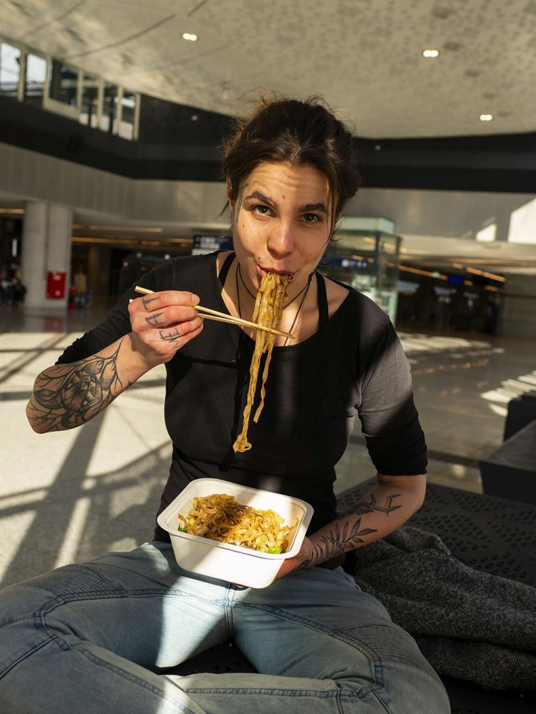Na zdjęciu widać kobietę siedzącą po turecku i jedzącą pałeczkami długi makaron