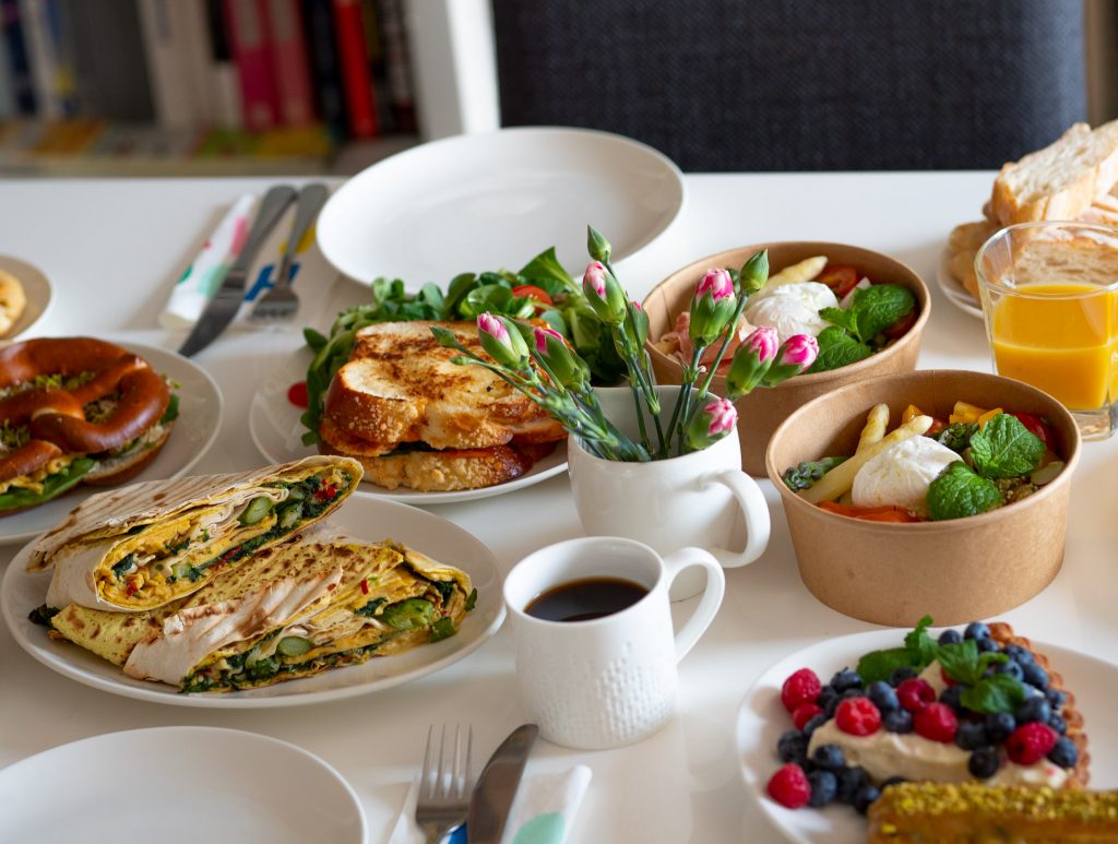 Na zdjęciu widać śniadanie ustawione na stole. Białe talerze i dania: chałkę, precel i tortillę. 
